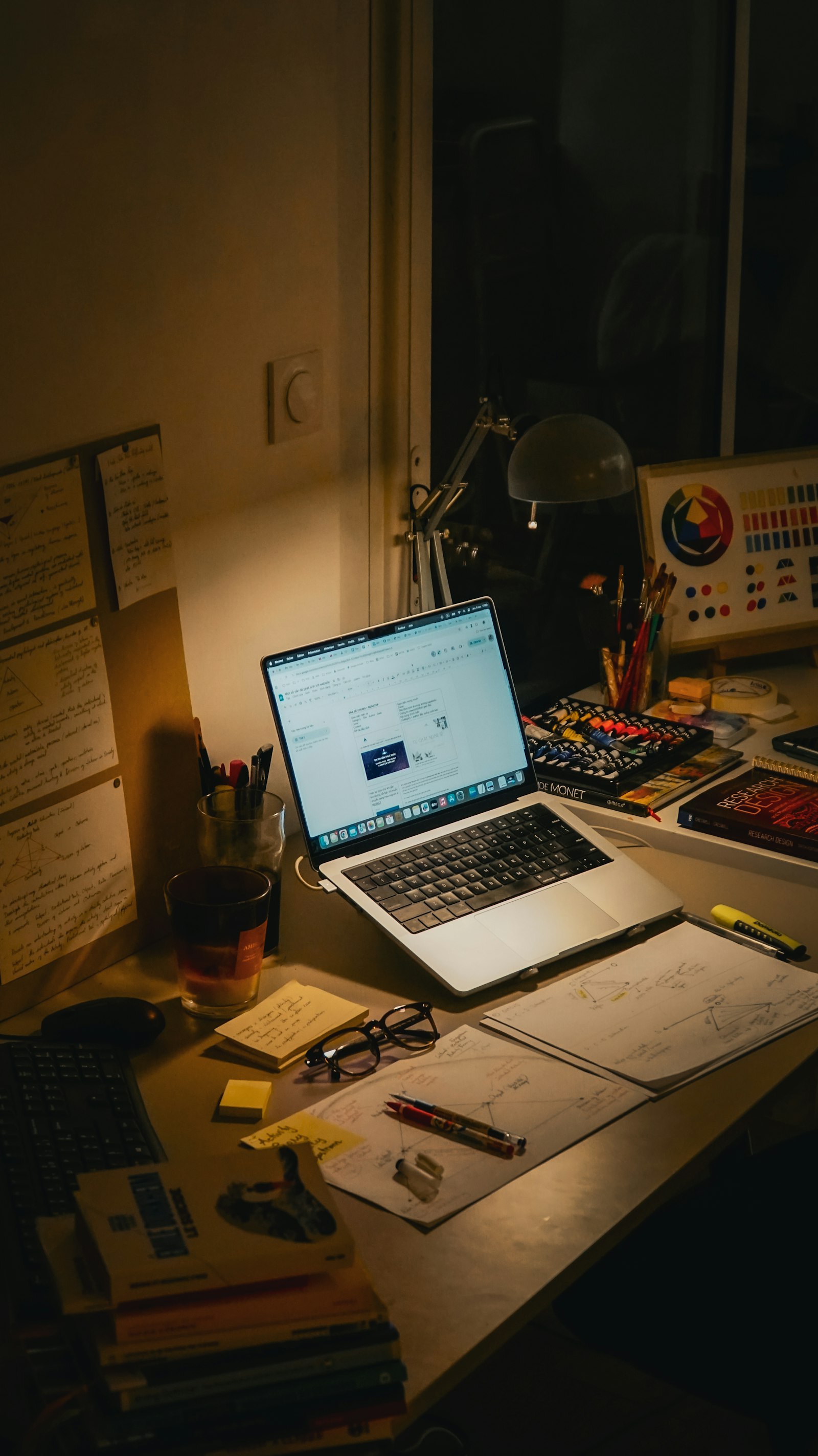A desk at night covered in handwritten notes, books, and a laptop
